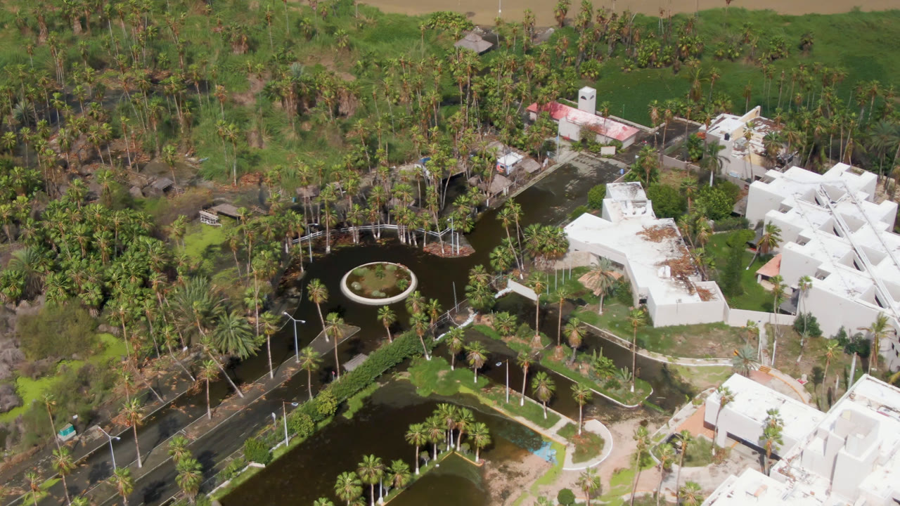 Aerial frame in San Jose del Cabo with the circular pond feature left of center, white low-rise buildings to the right, a road through the park, and no visible people.