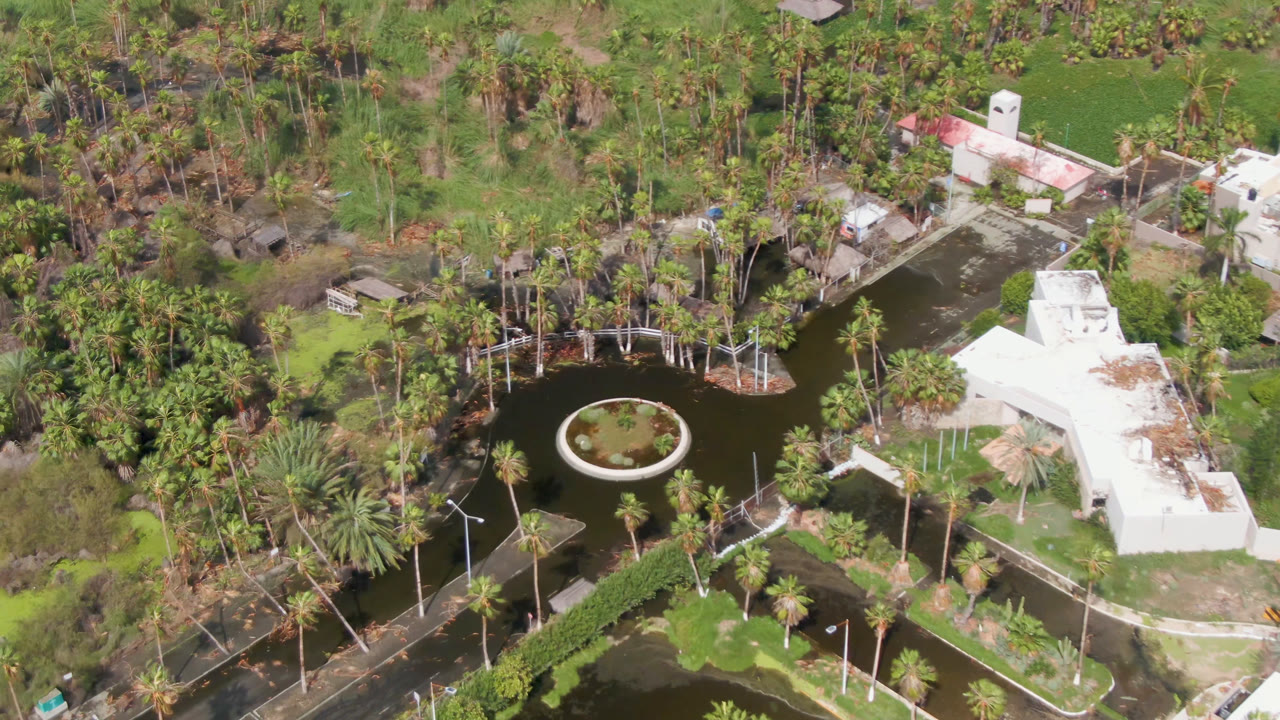Opening aerial frame in San Jose del Cabo shows a dark landscaped pond with a circular planted island, palms, green paths, small structures, and no visible people.