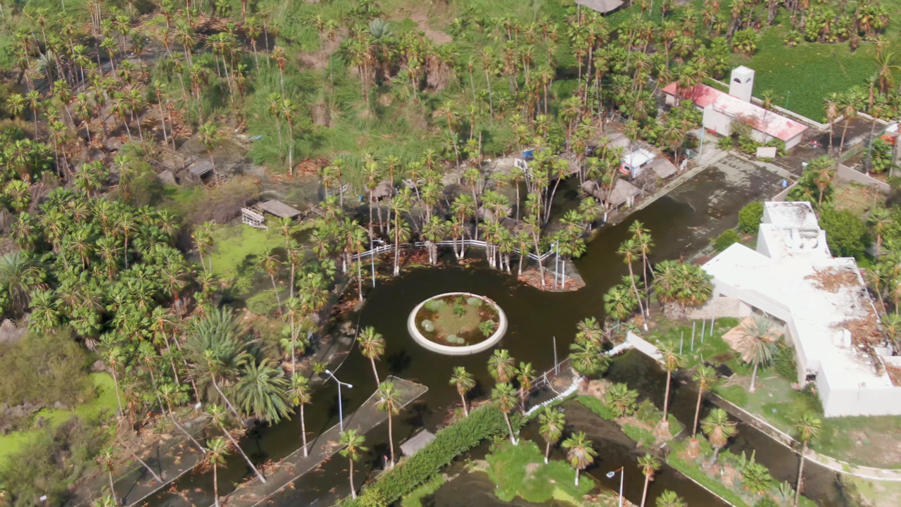 At one second, the San Jose del Cabo aerial view keeps the round island in the pond near center, with white roofs, dense palms, paths, and no visible people.