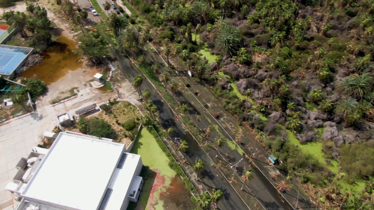 Clear overhead view of a white-roofed building, road, palms, pond water, and sport courts.