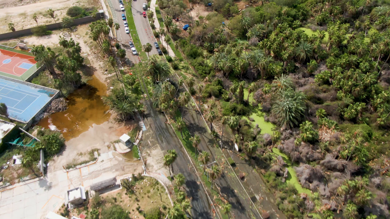Aerial view in San Jose del Cabo with the tennis courts more centered, brown water beside the courts, parked cars along the road, mixed palms, and no visible people.