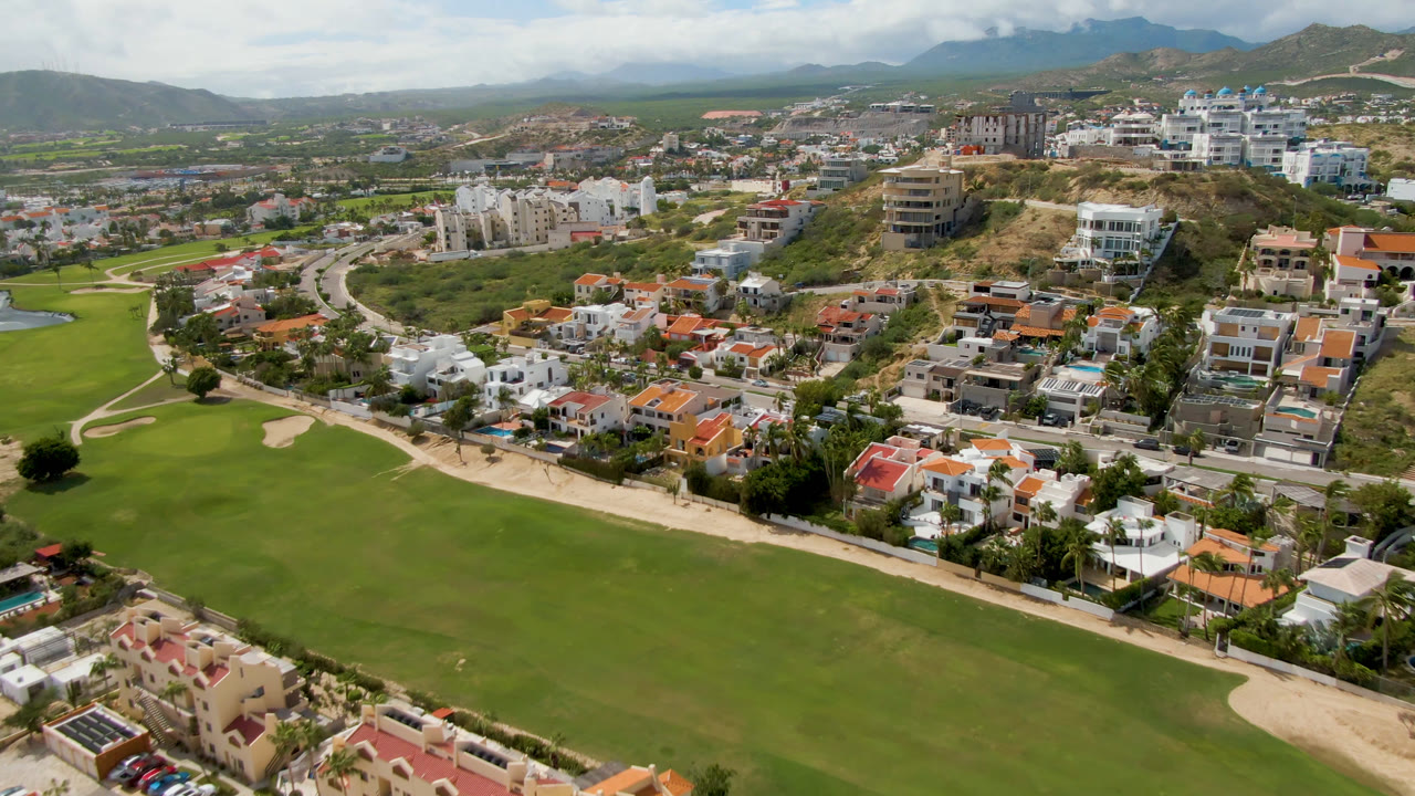 Aerial frame from San Jose del Cabo showing a broad green fairway, sand traps, residential buildings, dry hills, cloudy sky, and no visible people.