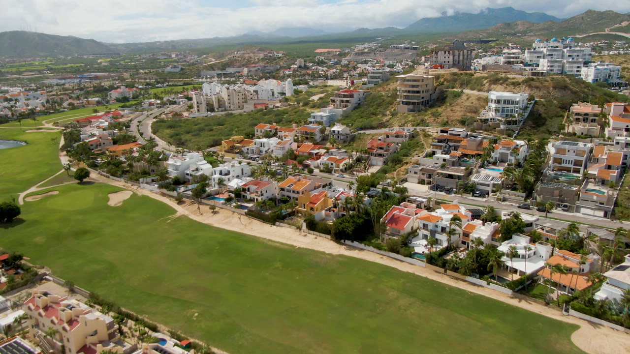 Aerial view in San Jose del Cabo with manicured fairway grass, pale bunkers, curved roads, hillside homes, distant mountains, and no visible people.