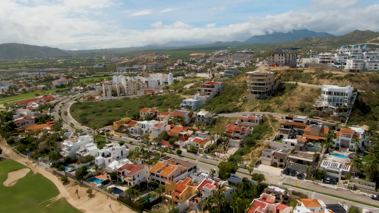 San Jose del Cabo aerial frame with a curving road through residential blocks, dry hillside lots, golf fairway at the lower edge, distant mountains, and no visible people.