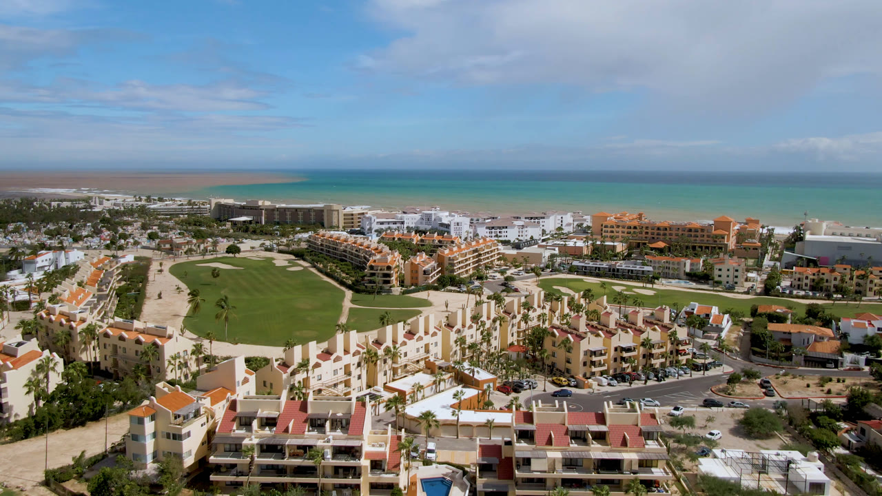 Wide aerial view over San Jose del Cabo resort buildings and a green golf course toward the beach and turquoise ocean, with no visible people.