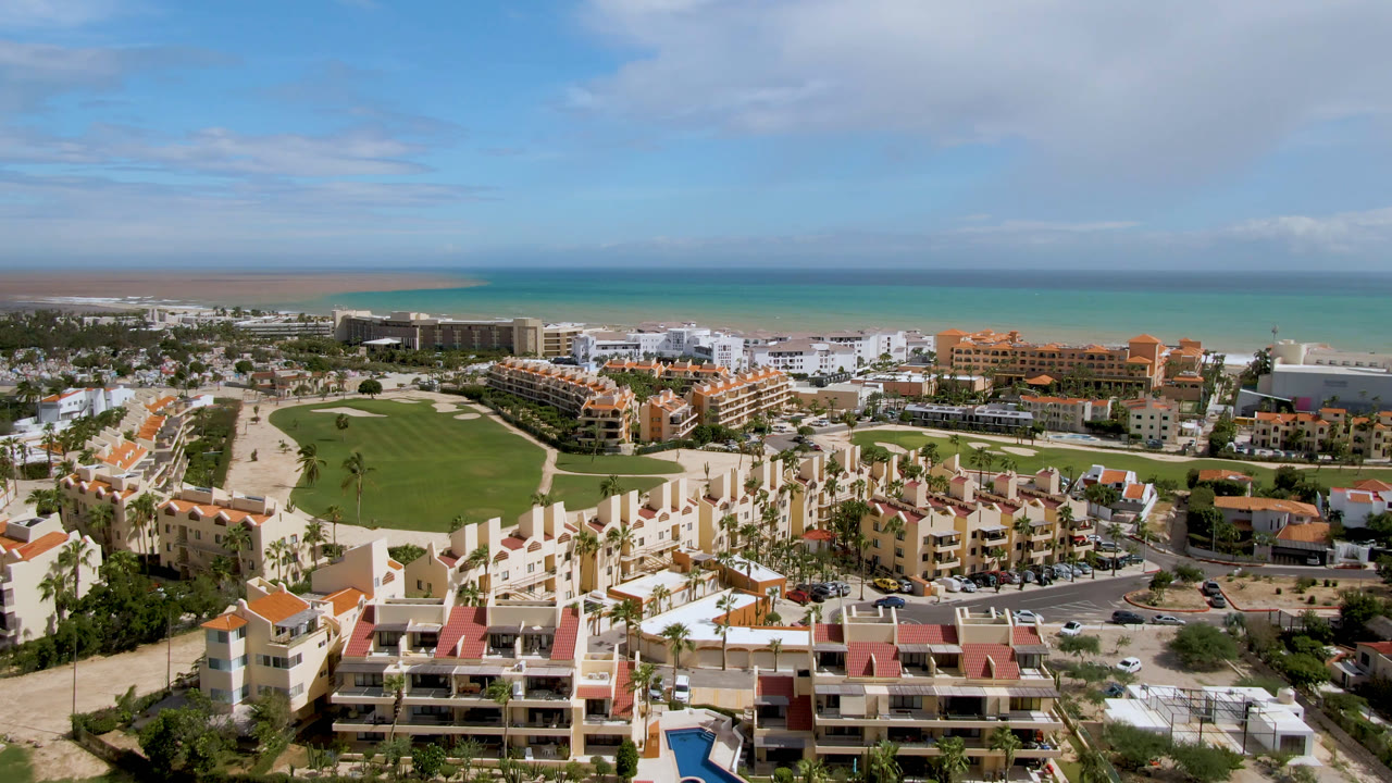 Elevated San Jose del Cabo view with cream resort buildings in front, a broad green fairway behind them, beachfront hotels, and a flat ocean horizon, with no visible people.