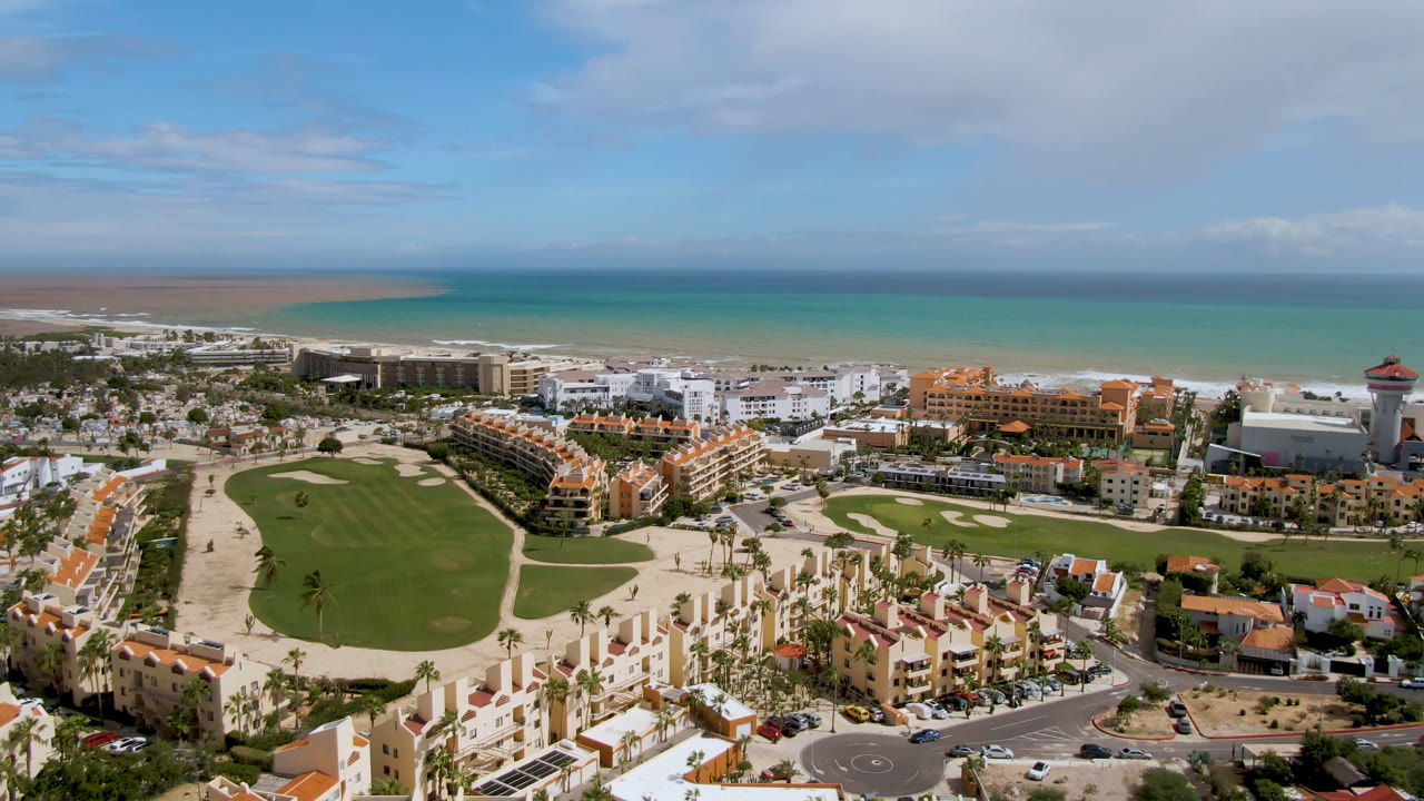 Aerial view of coastal buildings, golf course, beach, and ocean under a partly cloudy sky.