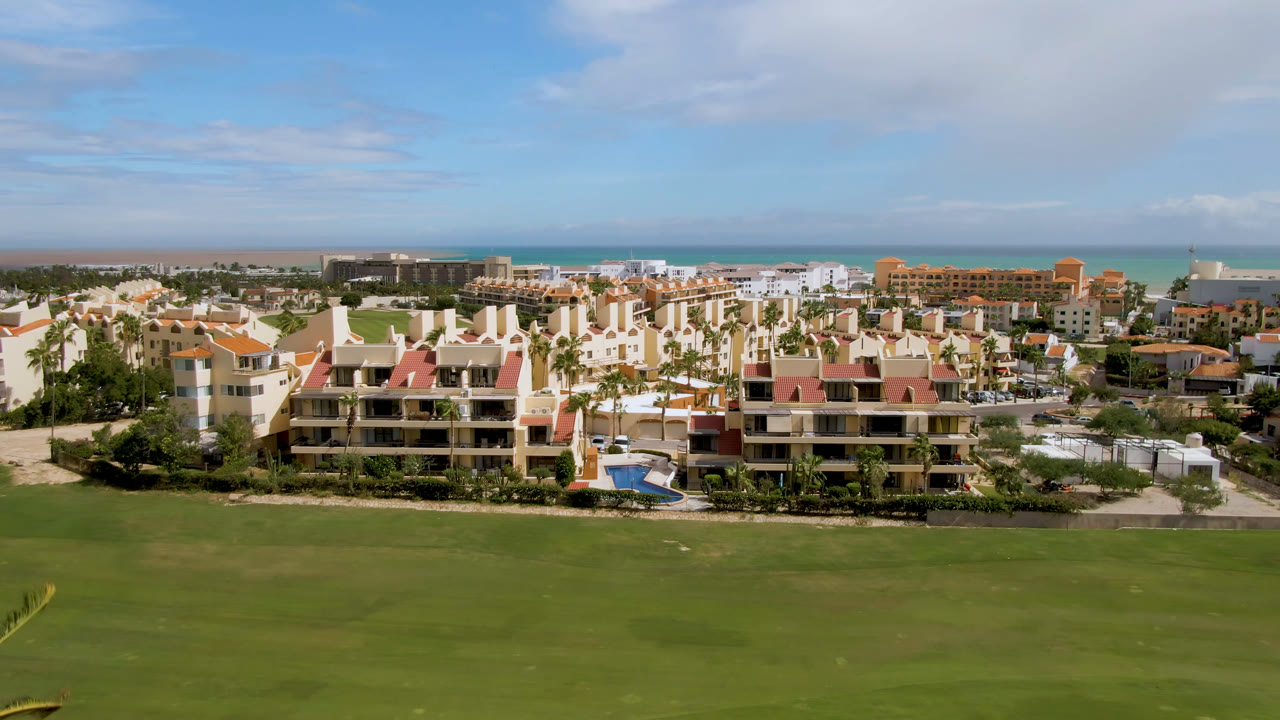 San Jose del Cabo residential blocks sit beyond a wide green fairway, with palm trees, the ocean horizon, scattered clouds, and no visible people.