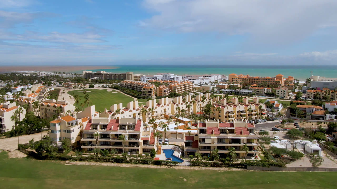 Opening aerial frame over San Jose del Cabo resort buildings, a green golf course in the foreground, turquoise ocean on the horizon, and no visible people.