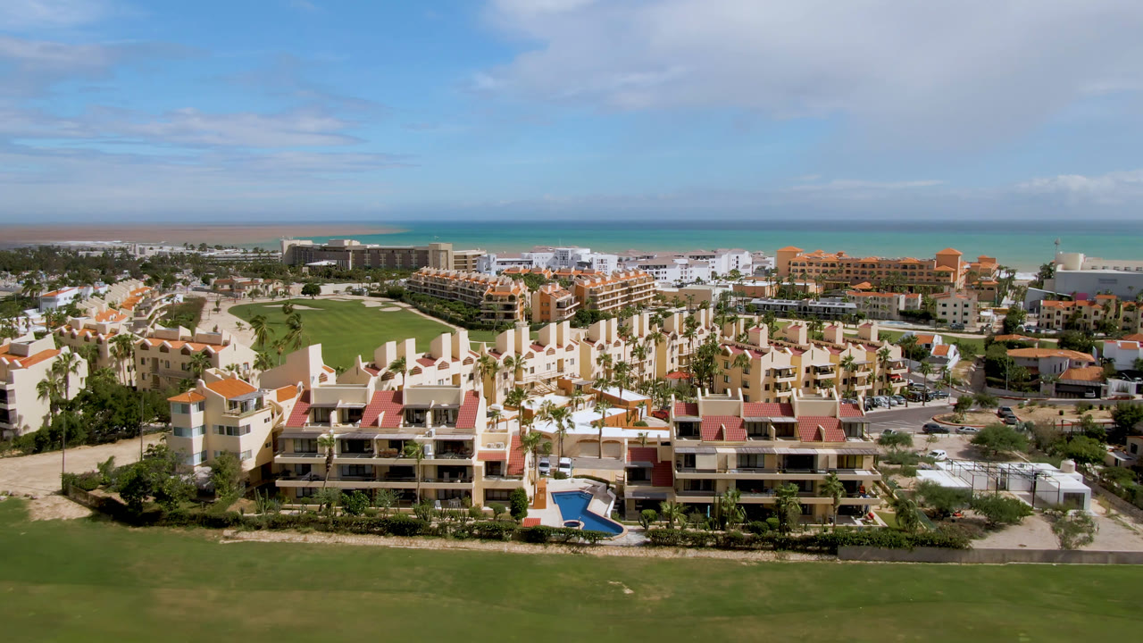 Aerial frame in San Jose del Cabo with beachfront resort blocks centered behind golf greens, palm-lined grounds, ocean beyond, and no visible people.
