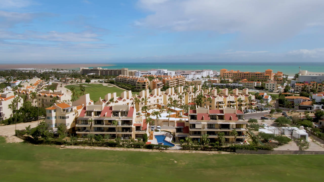 Aerial view over San Jose del Cabo showing red-roofed resort buildings, bright green fairway, beach line, blue-green ocean, and no visible people.