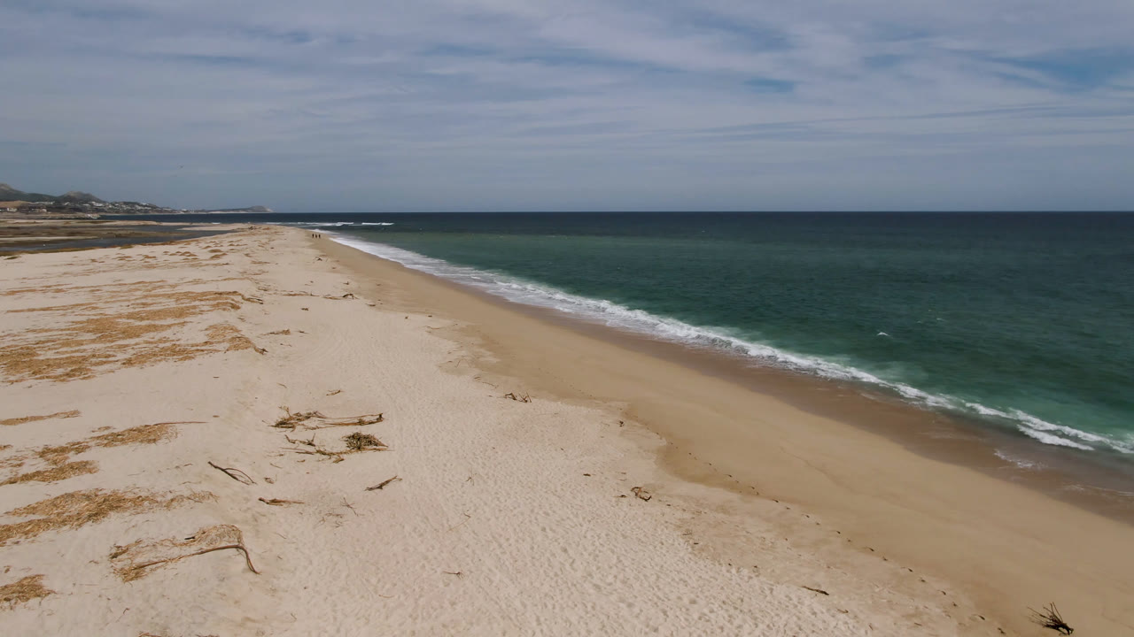 Opening aerial frame over a sandy San Jose del Cabo beach with scattered driftwood, green-blue ocean to the right, white surf along the shore, and no visible people.