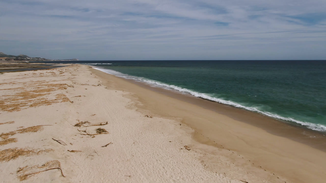Aerial view along the San Jose del Cabo shoreline with pale sand in the foreground, small debris marks across the beach, ocean on the right, and no visible people.