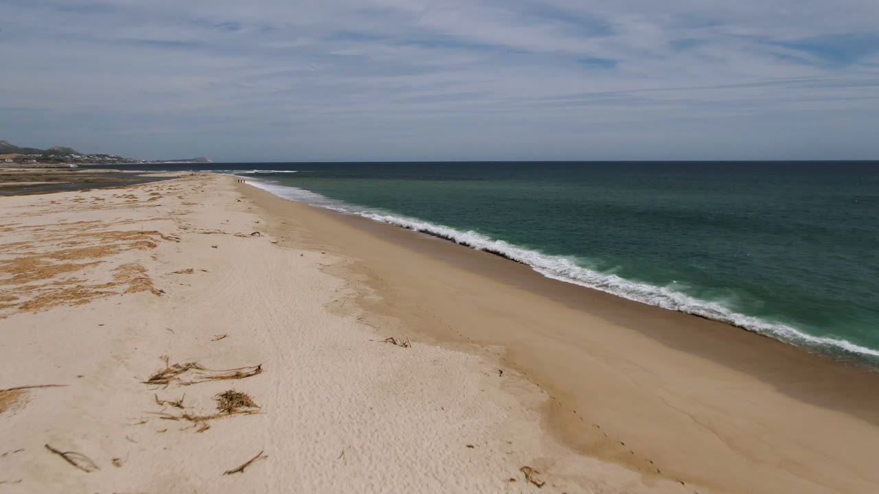 San Jose del Cabo aerial shoreline frame with the surf line closer to the right edge, a broad stretch of light sand, distant coast at left, layered clouds, and no visible people.