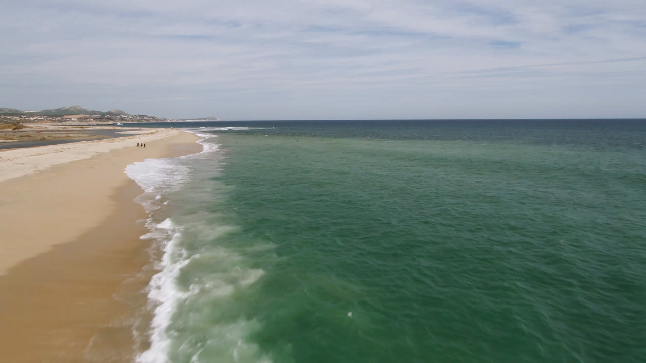Aerial view of sandy beach, surf, green ocean, cloudy sky, and tiny beach walkers.