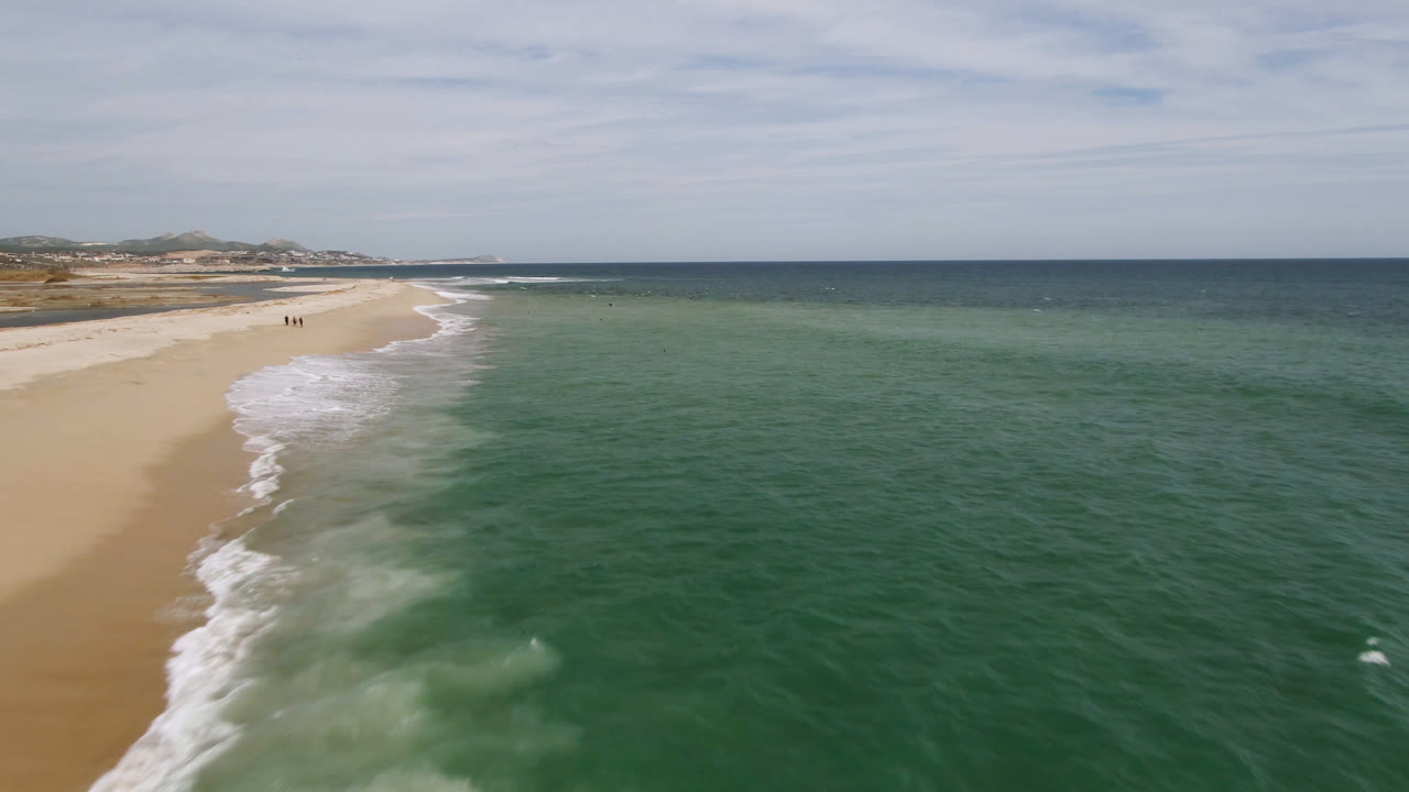 A wide aerial frame of San Jose del Cabo coastline shows more open ocean to the right, foamy waves curling onto the sand, and small distant people walking near the upper left beach.