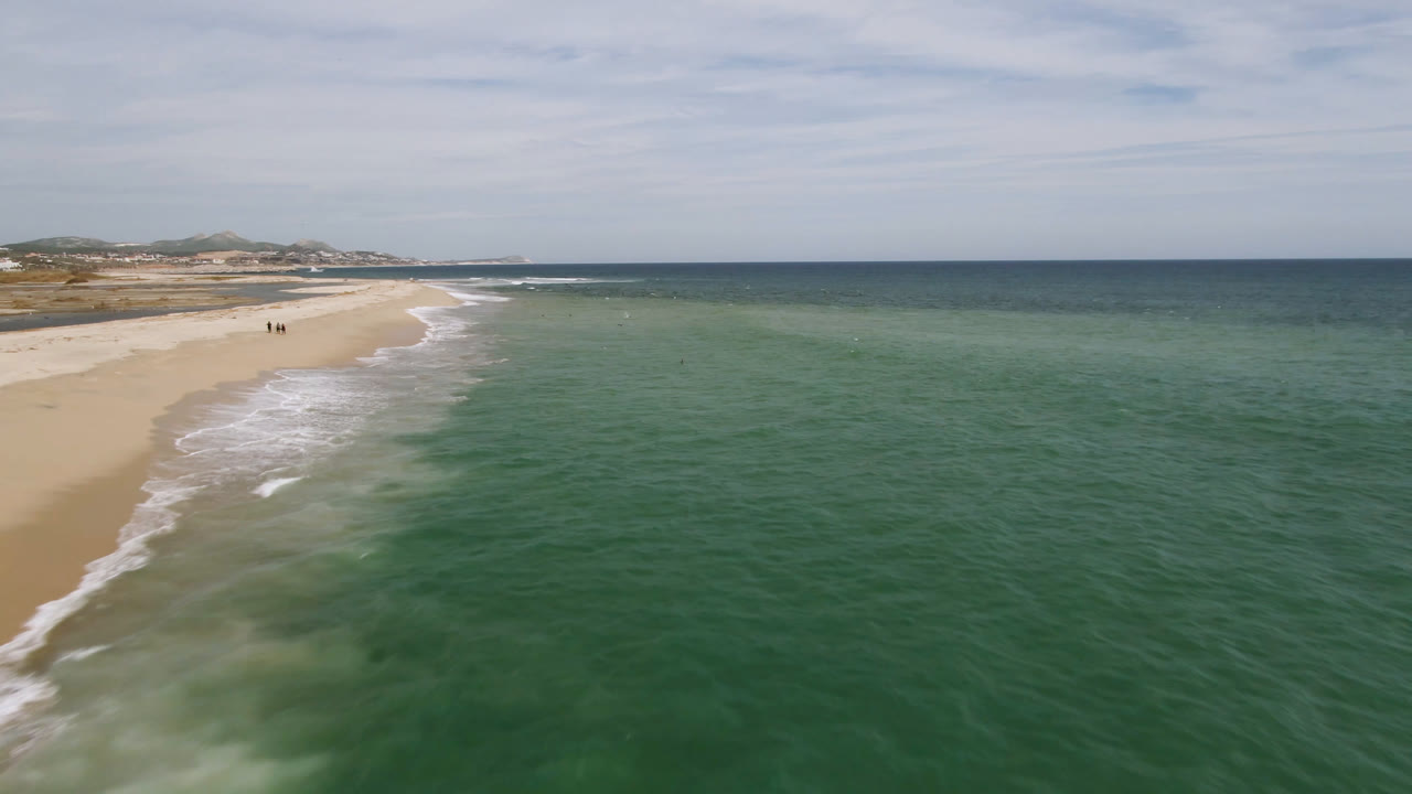San Jose del Cabo shoreline appears from above with the beach receding into the distance, green water filling most of the frame, and a few tiny people visible on the sand.