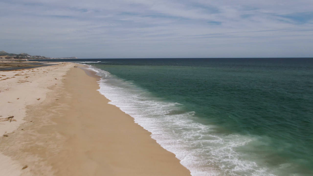 Aerial frame over San Jose del Cabo beach with sand on the left, green-blue ocean on the right, white surf tracing the shoreline, distant coast, and no visible people.