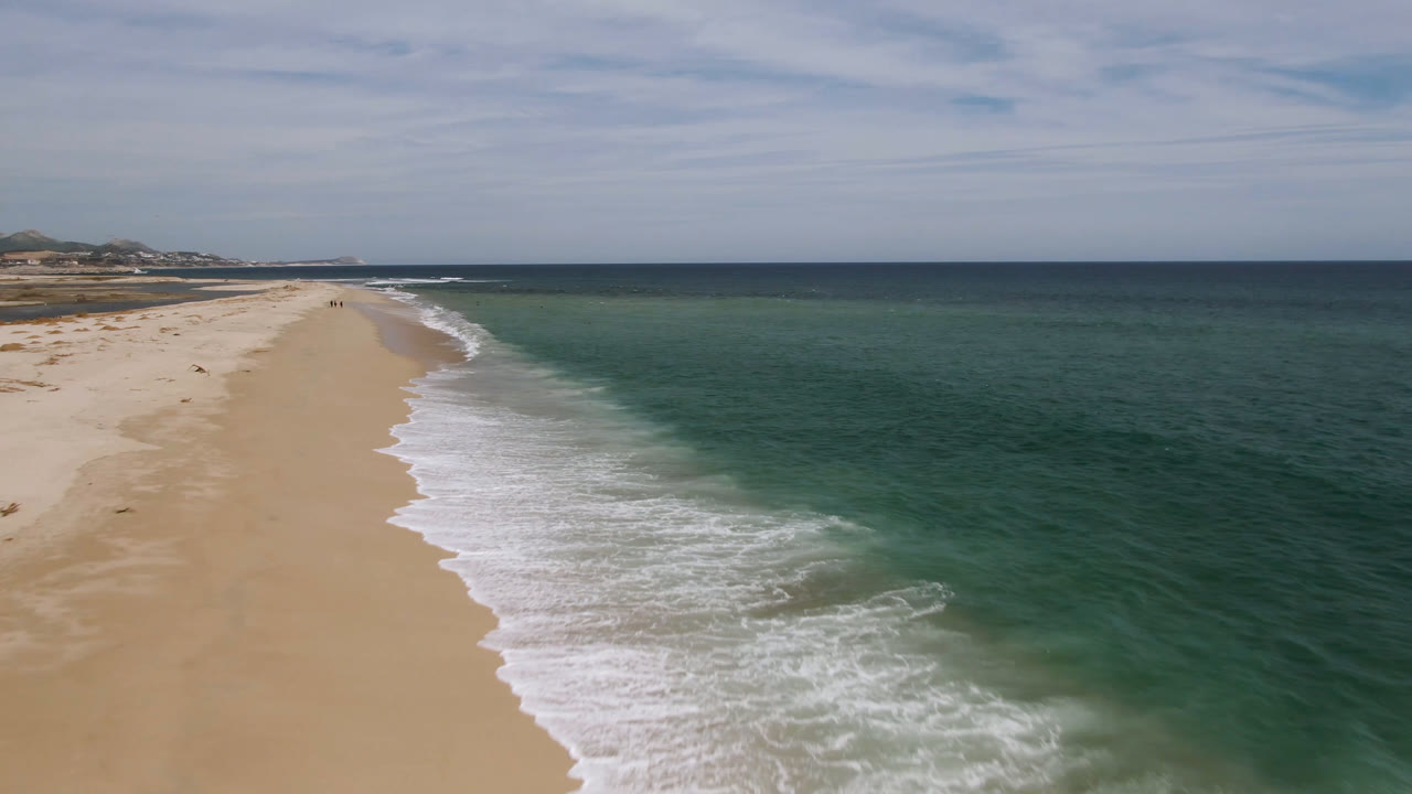 Drone view over San Jose del Cabo shoreline with a wider band of foamy surf advancing up the sand, open ocean to the right, pale clouds overhead, and no visible people.