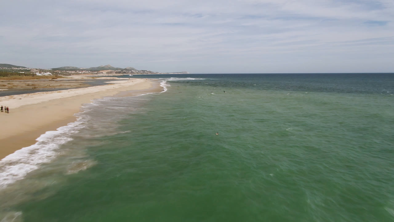 Aerial frame over San Jose del Cabo shoreline, with white surf tracing the sand at left and open green water stretching to the horizon, no visible people.
