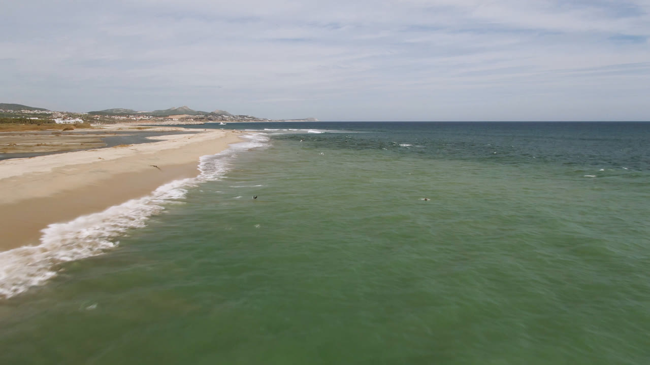 Wide aerial view of green-blue ocean meeting a pale sand beach under clouds.