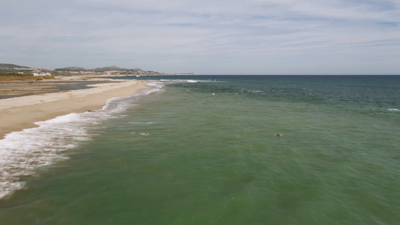 Aerial view over San Jose del Cabo beach with the surf line pushing diagonally from the sand into green water, low hills beyond the shore, and no visible people.