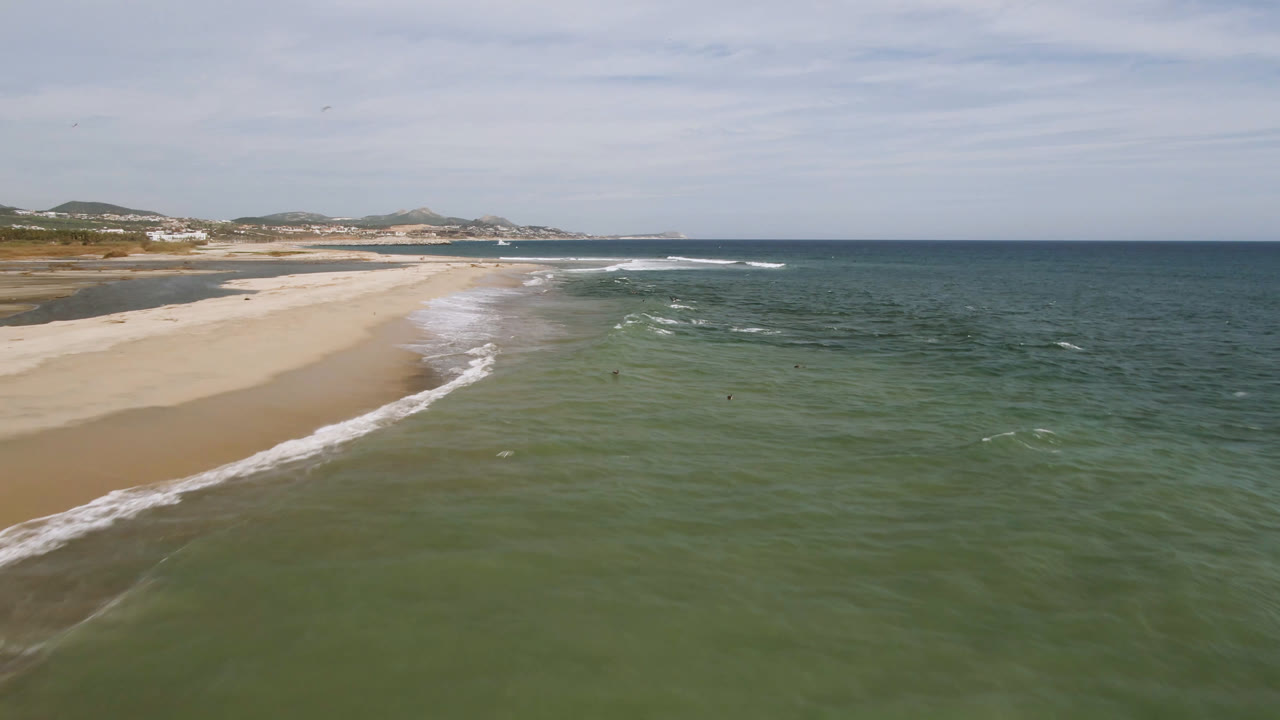 Aerial view of San Jose del Cabo beach as a white wave band advances along the sand, with darker ocean water offshore, distant shoreline development, and tiny possible swimmers in the water.