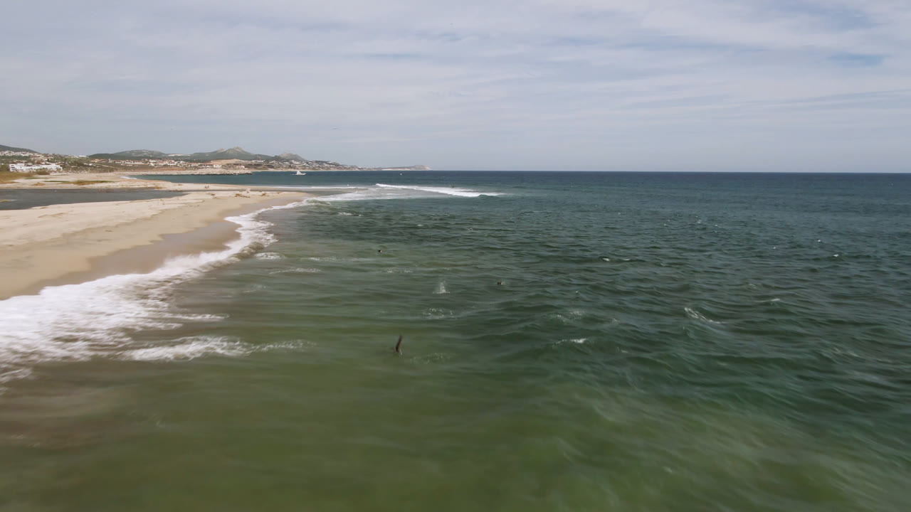 Aerial view over the San Jose del Cabo shoreline with green-blue ocean in front, a sandy beach at left, distant hills beyond the coast, and tiny surfers too small to identify.