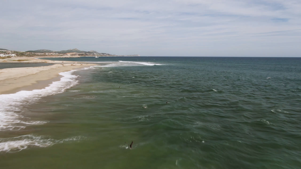 Drone view of the San Jose del Cabo coast as white water reaches the beach at left and small dark surfer shapes sit in the green water near the lower center.