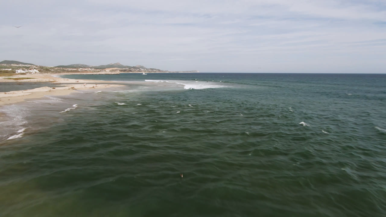 Opening aerial frame over the San Jose del Cabo coastline, with dark green water in front, a sandy beach on the left, low hills beyond, thin clouds overhead, and no visible people.