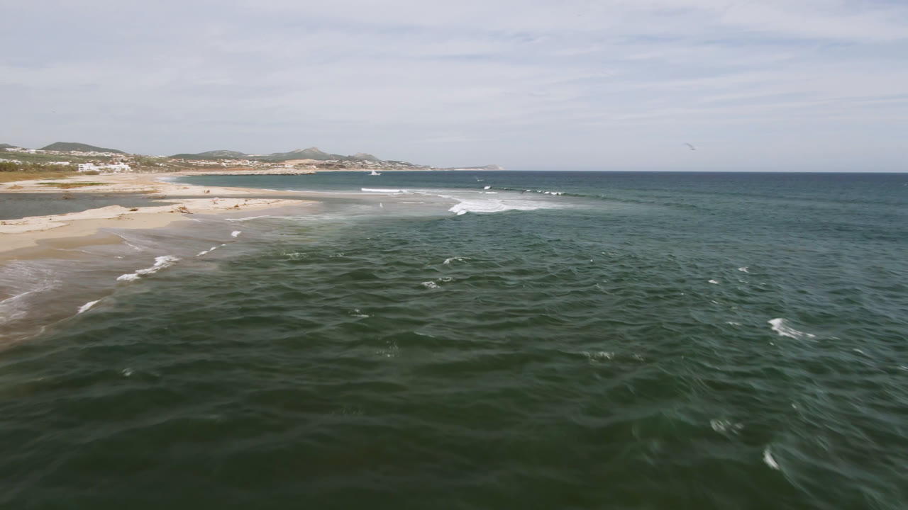 San Jose del Cabo beach view one second in, showing choppy green-blue water, small white wave lines near shore, sand stretching left, distant coastal buildings, and no visible people.