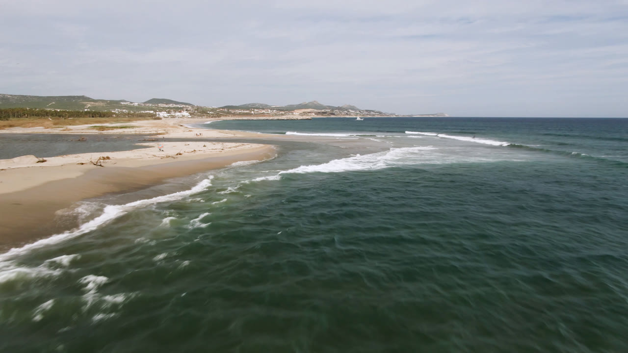 Aerial frame over San Jose del Cabo coastline with green-blue water in the foreground, white surf meeting a curved sandbar, distant hills and buildings, and no visible people.