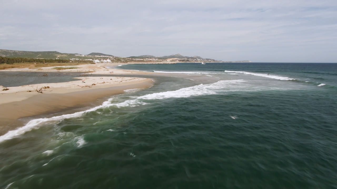 Aerial view in San Jose del Cabo with the surf band slightly farther across the frame, tan beach on the left, open ocean on the right, distant hills, and no visible people.