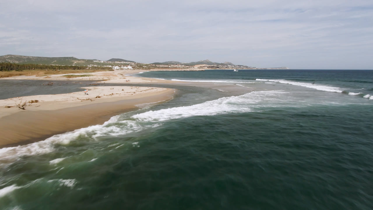 Aerial coastal view with a foamy wave crossing dark green water beside sand.