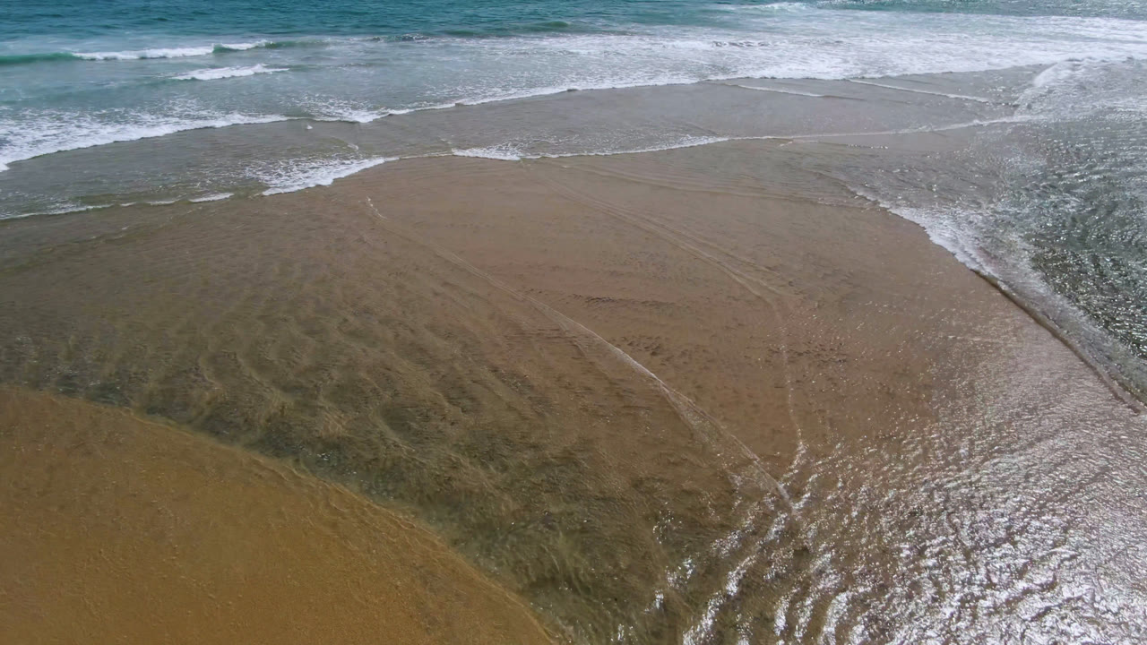 Aerial view in San Jose del Cabo, Baja California Sur, with white surf spreading diagonally across wet tan sand and no visible people.