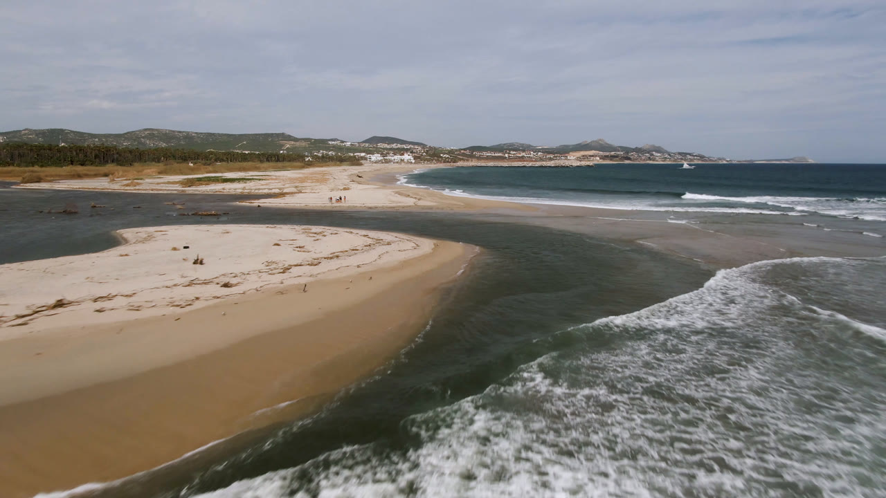 Aerial view in San Jose del Cabo with the sandbar more centered, surf breaking at right, low dunes at left, distant shoreline, and tiny distant beachgoers on the sand.
