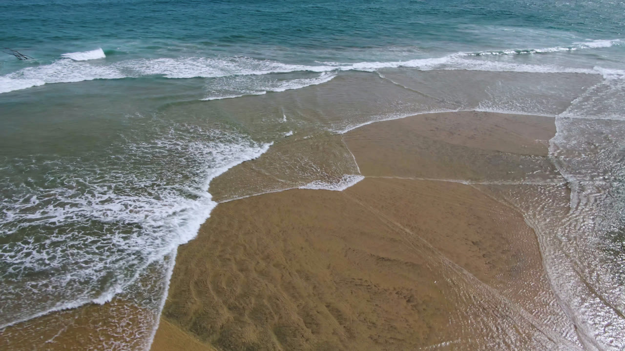 A white wave edge curls diagonally over shallow water and exposed sand in San Jose del Cabo, with no visible people.
