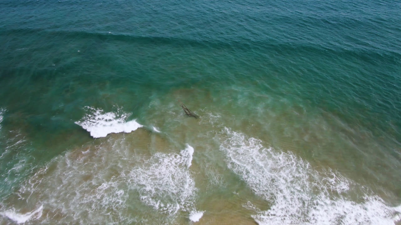 Overhead view in San Jose del Cabo, Mexico, showing green-blue coastal water, breaking white foam, sandy shallows, and a small dark surfer or board-like shape near the upper wave line with no visible faces.