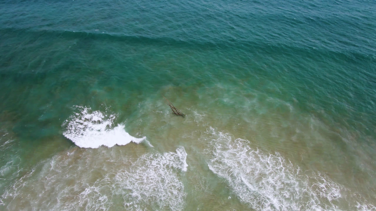 A later overhead frame from San Jose del Cabo shows white foam widening across turquoise shallows while the small dark surfer or board-like shape sits slightly right of the main break, with no visible faces.