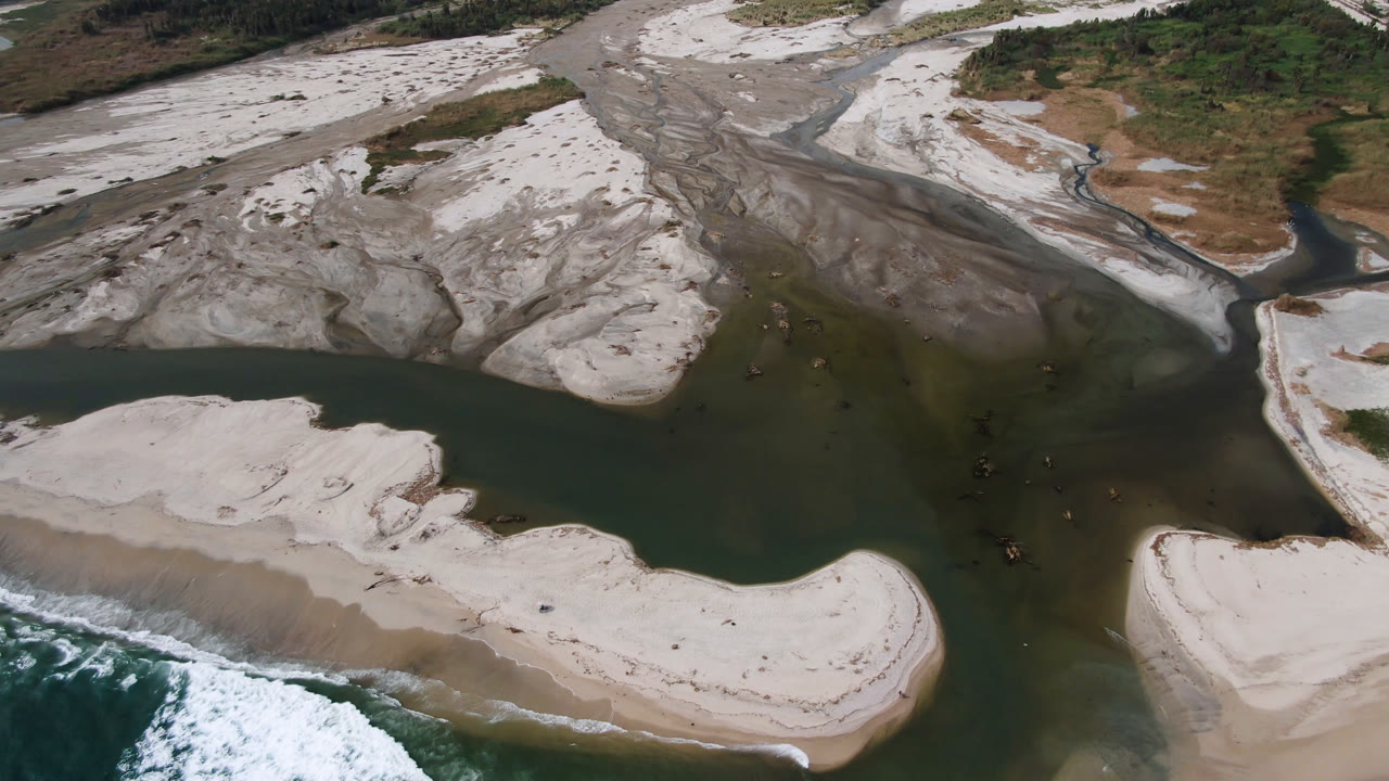 Aerial frame in San Jose del Cabo with a broad dark water channel, curved light sand bars, white surf entering from the bottom edge, and no visible people.
