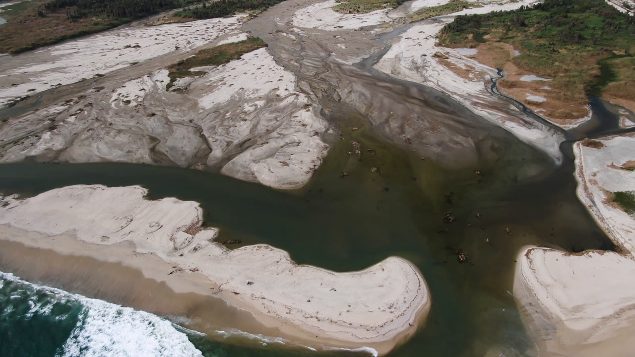 Drone-like overhead view of a river mouth, sand flats, and surf on a beach.