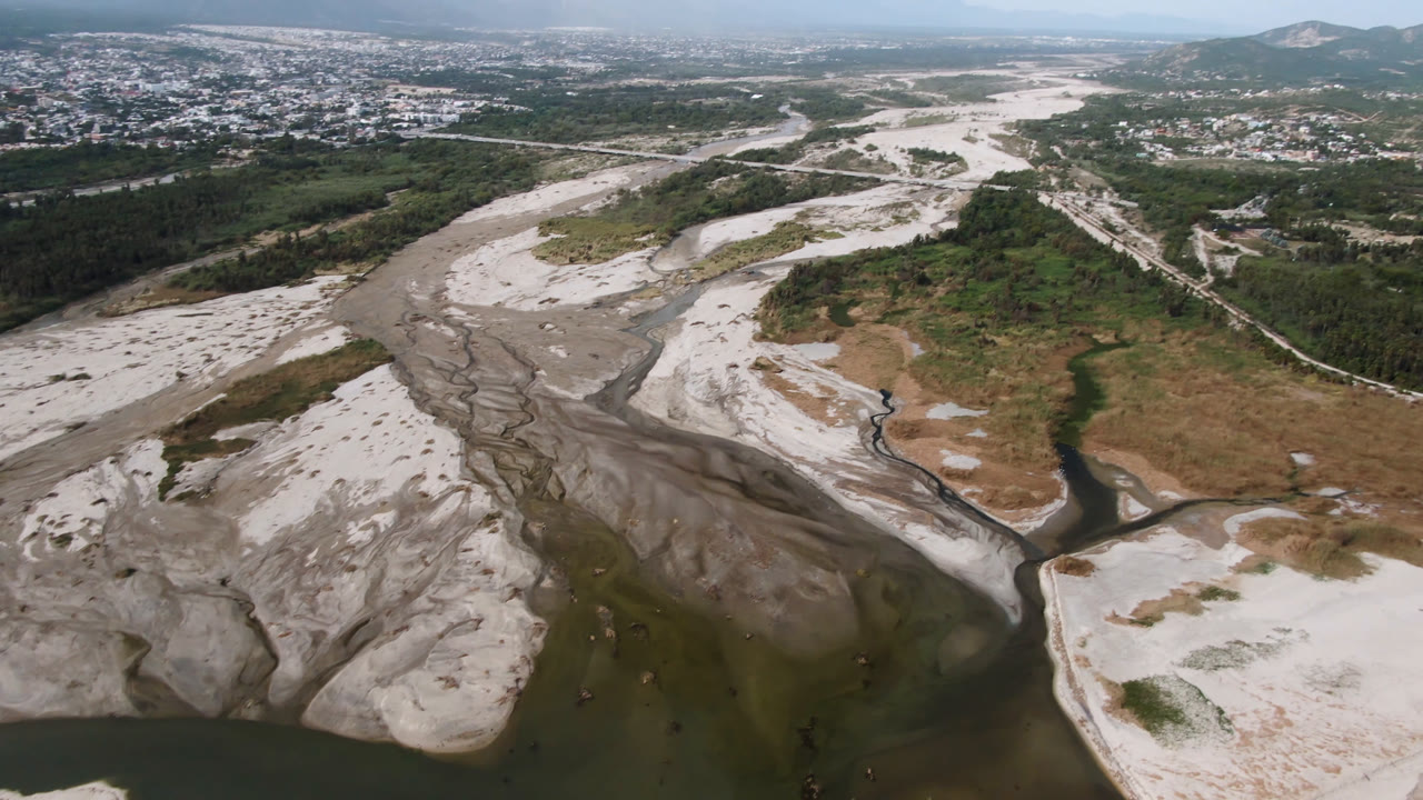 Aerial view over a dry riverbed in San Jose del Cabo, Mexico, with pale sand bars, dark branching water channels, green vegetation, distant hills, and no visible people.