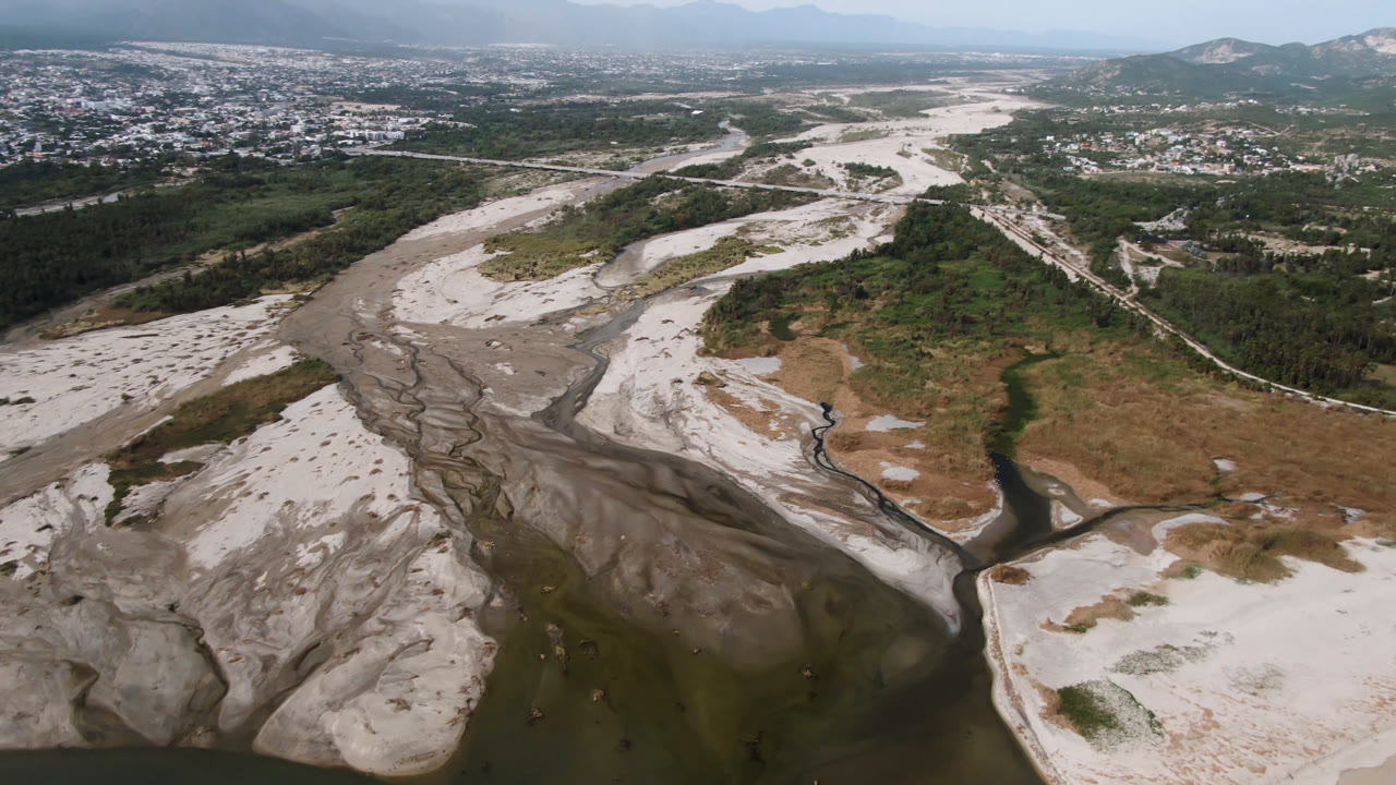 Wide aerial frame in San Jose del Cabo, Mexico, looking across pale river sand, braided dark channels, a green valley strip, distant urban development, hills, and no visible people.