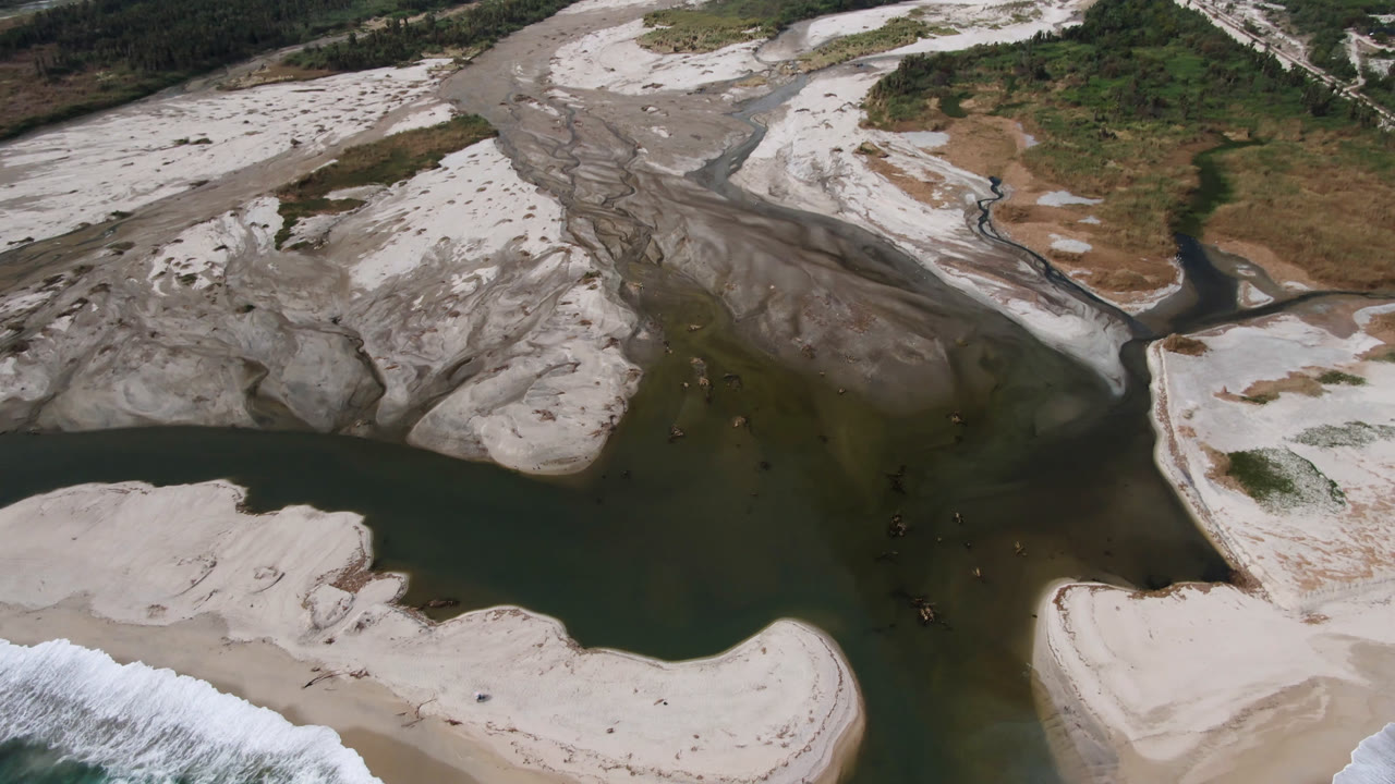 Aerial frame in San Jose del Cabo showing dark river water meeting pale sand bars with surf entering at the lower left, no visible people.