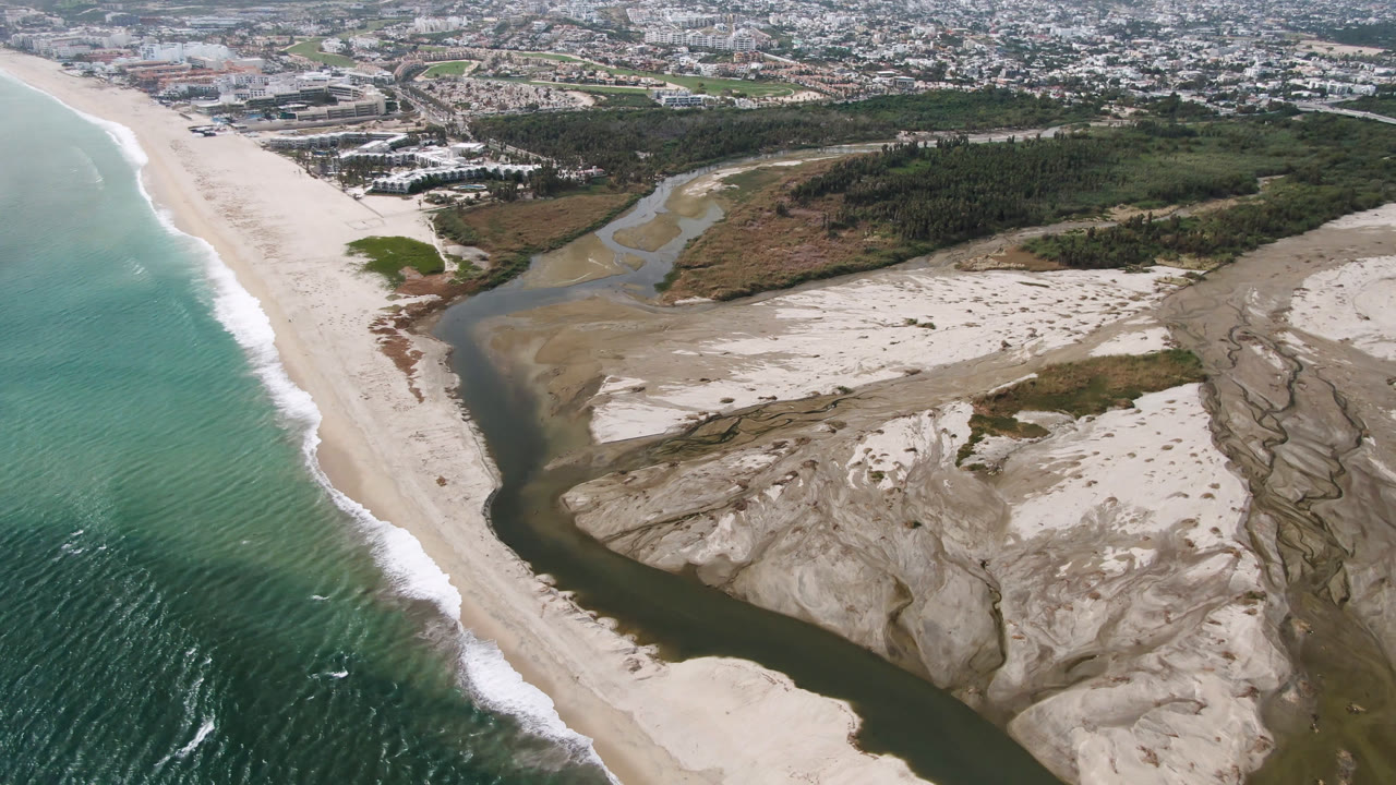 Aerial frame over San Jose del Cabo showing teal ocean beside a pale beach, a dark river channel cutting through sand banks, town buildings at the top, and no visible people.