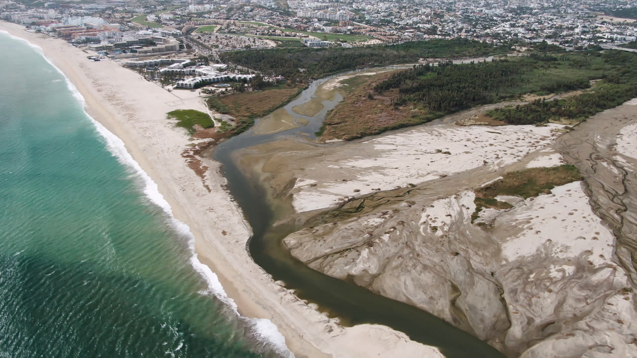 Aerial view of ocean, beach, river channel, sand banks, vegetation, and distant buildings.