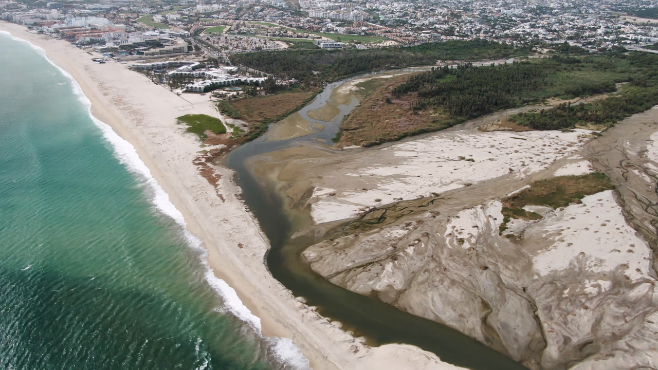 Aerial frame in San Jose del Cabo with more surf texture visible on the left, the beach running diagonally, river water crossing the sand banks, scrubland beyond, and no visible people.