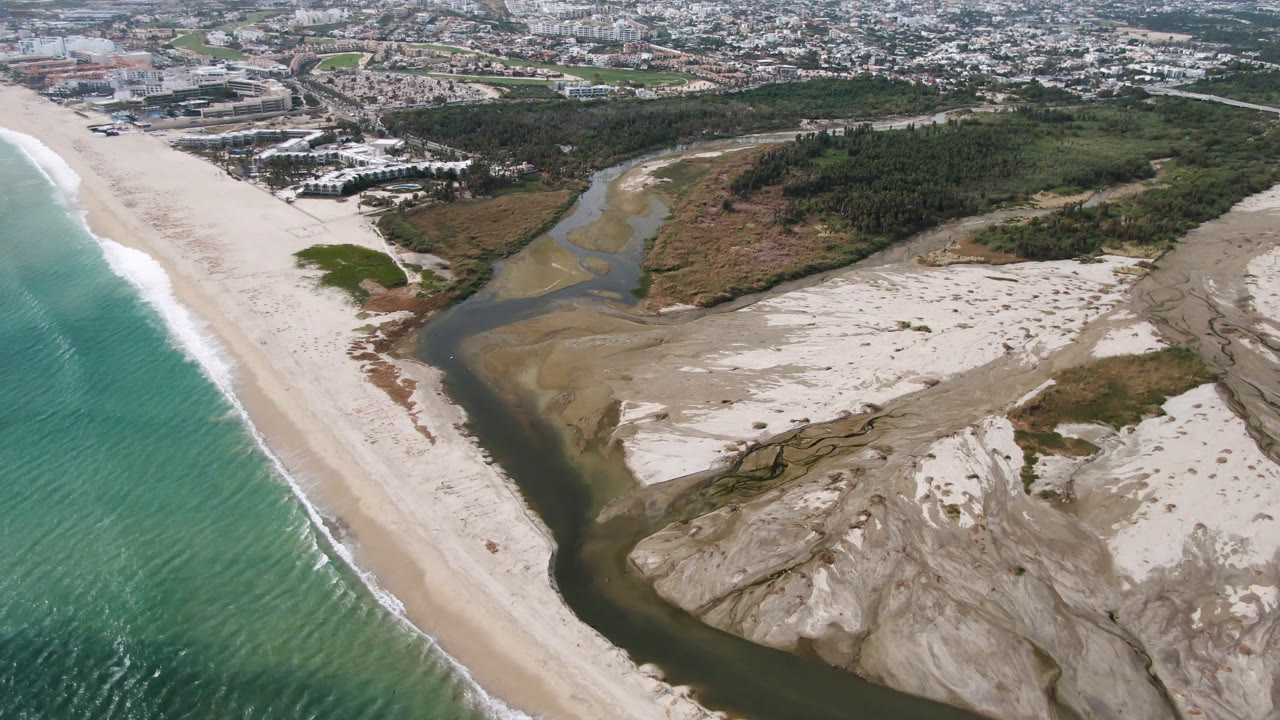 Aerial view over San Jose del Cabo showing turquoise surf beside a wide pale beach, a dark river channel through sand banks, scrub vegetation, distant buildings, and no visible people.