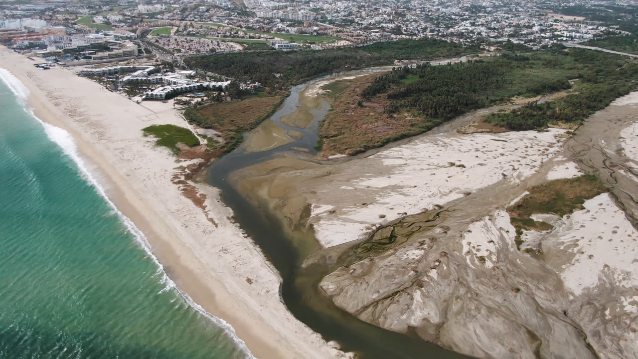 Drone frame in San Jose del Cabo with the river channel centered between pale sand flats, the ocean surf running along the left beach edge, distant low buildings, and no visible people.