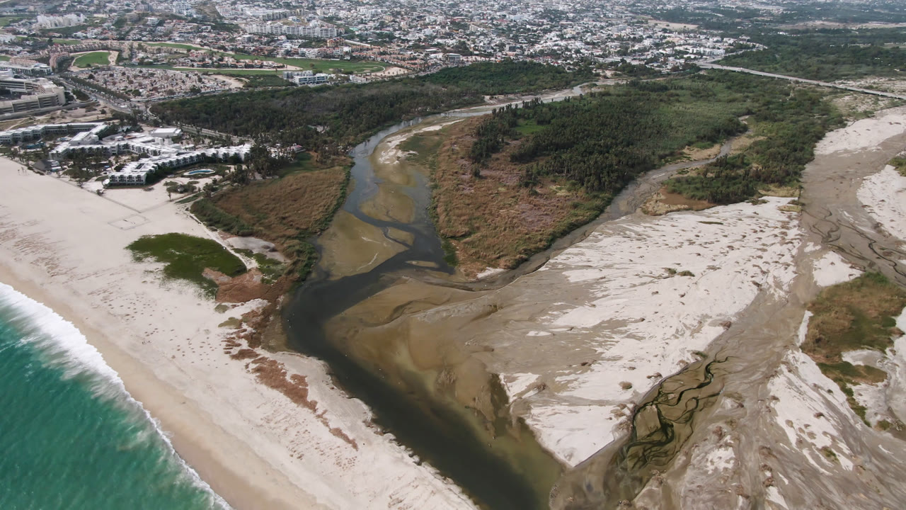 Aerial view over San Jose del Cabo, Mexico, where a dark river channel spreads across pale beach sand toward turquoise surf, with vegetation and town blocks inland and no visible people.